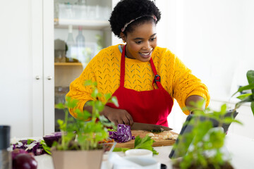 Happy plus size african american woman in apron preparing meal using tablet in kitchen
