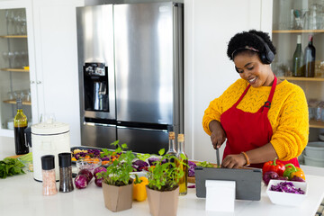 Plus size african american woman in apron and with headphones preparing meal using tablet in kitchen