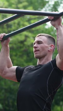 Slow motion handheld shot of powerful muscular sportsman listening to music on earbuds and doing pull ups on horizontal ladder during intense outdoor training Vertical video