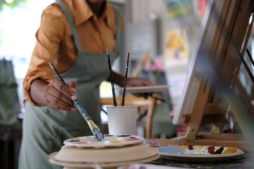 Hapy young curly art school female student painting picture with brush on canvas in art studio