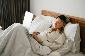 A young girl, of European appearance with blond hair, works remotely on a laptop, lying in bed under a blanket on a pillow.