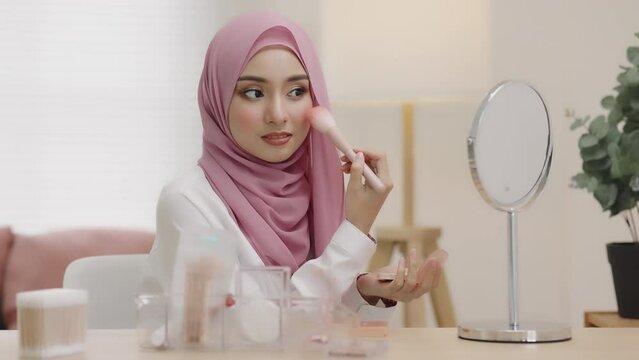 Young asian beautiful muslim woman with hijab looking mirror applying makeup with brusher of cheek daily routine at home. Beautiful of girl holding brush make-up on face.