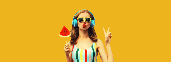 Summer portrait of stylish woman in headphones listening to music blowing her lips with juicy lollipop or ice cream shaped slice watermelon on yellow background