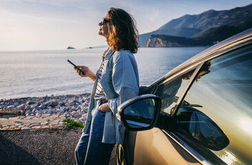 Young woman traveler istanding by her car during summer sea holiday on the beach at sunset - obrazy, fototapety, plakaty