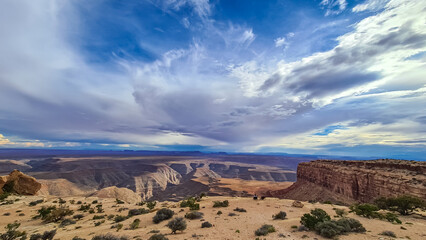 Naklejka premium Woman sitting in camping chair enjoying scenic aerial vistas of desert landscape of Valley of the Gods seen from Muley Point near Mexican Hat, San Juan county, Utah, USA. Monument valley in distance