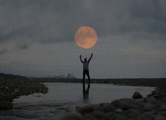 Silhouette of man with hands up against the background of the sunset in the reflection of the sea or pond.  Man Raising His Hands or Open arms.  down town on background.  full moon above head