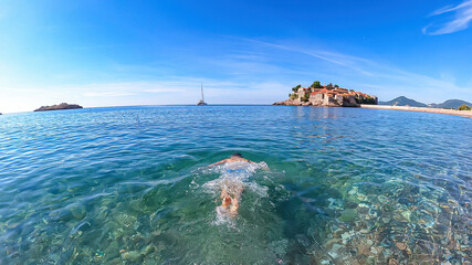 Man bathing in crystal clear water at sand beach next to idyllic island Sveti Stefan, Budva Riviera, Adriatic Mediterranean Sea, Montenegro, Europe. Summer vacation in luxury hotel resort at seaside