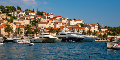 Panoramic view of croatian holiday hot spot old village of Hvar on Hvar island in the Adriatic Sea. Waterfront promenade with motor yachts and fisher boats on a sunny summer day with warm light. - obrazy, fototapety, plakaty