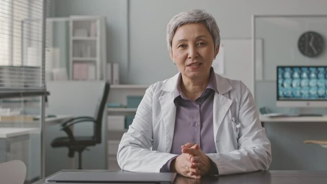 Waist up POV of mature Asian female doctor sitting at workplace in her office looking at camera and giving consultation to patient during appointment