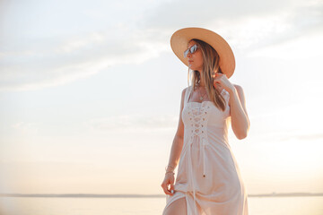 Young caucasian woman in white dress, summer hat and sunglasses posing on beach on sunset