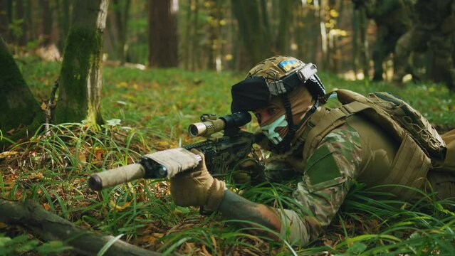 Footage of young Caucasian man in camouflage clothes and balaclava lying on grass, holding weapon and aiming target during tactical action in autumn forest. Concept of war and attacking. Outdoors