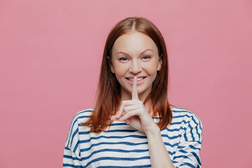 Pleased woman, brown hair, gentle smile, finger on lips. Silence gesture. Striped clothes. Pink background