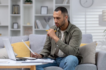 Worried and serious young African American man working at home with documents and bills. He sits huddled on the sofa in front of the laptop and holds papers in his hands