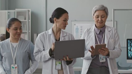 Three multiethnic female doctors discussing work while walking together along office with laptop and digital tablet in hands - Powered by Adobe