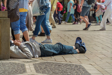 Authentic tramp sleeping on a stone-paved street on a sunny summer day against the background of a large group of passers-by and tourists, people walk past a needy man lying on the city street