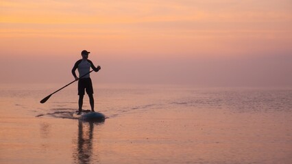 Man paddling on stand up paddle board against background of rising sun. SUP