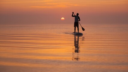 Man paddling on stand up paddle board against background of rising sun. SUP