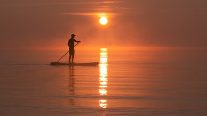 Man paddling on stand up paddle board against background of rising sun. SUP