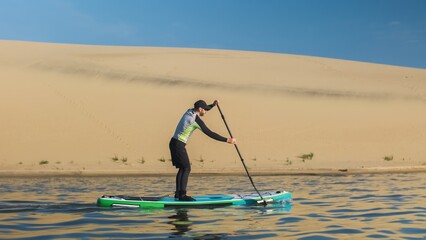 Man paddling on stand up paddle board against background of dunes in desert. SUP