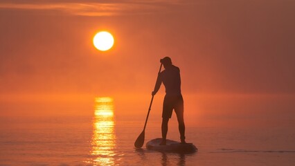 Man paddling on stand up paddle board against background of rising sun. SUP