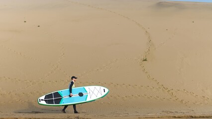 Man walking on shore with stand up paddle board along big dunes in desert. SUP
