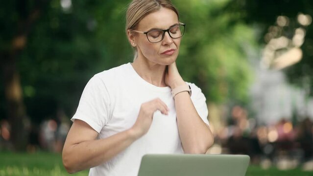 Overworked tired blond mature woman freelancer with severe neck shoulder pain after hard work on laptop computer outdoors Sad female suffers from tension in the spine at green park