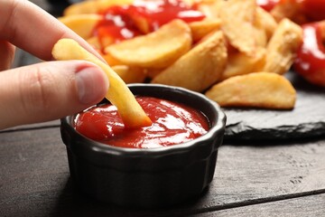 Woman dipping french fry into bowl with tasty ketchup at dark wooden table, closeup