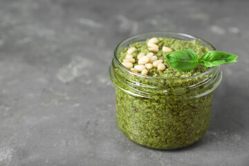 Jar with delicious pesto sauce, pine nuts and basil leaves on grey table, closeup. Space for text
