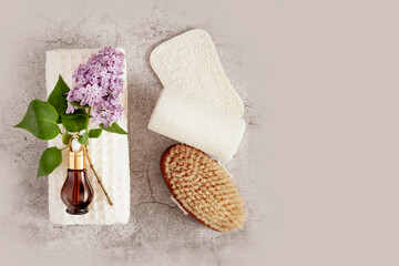 Spa treatment flat lay with bottle of organic oil essence collagen. Dropper bottle with natural serum on a white towel with lilac flowers, massage brush and loofahs on grey background. Copy space