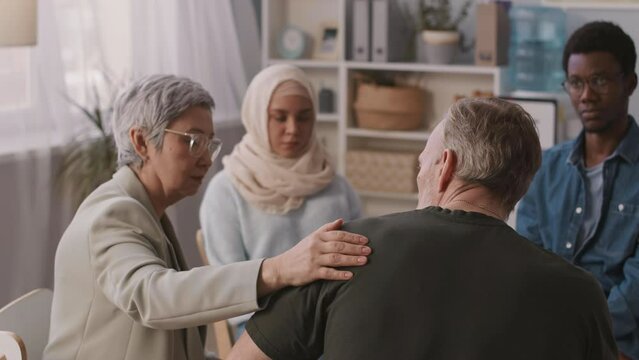 Back view of Asian female psychologist putting her hand on shoulder of depressed adult man attending group therapy sessions with other patients
