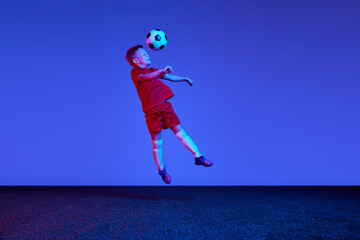 Fototapeta na wymiar One sportive child, boy in red uniform playing, training football over dark blue background in neon. Concept of action, sportive lifestyle, team game, health, energy