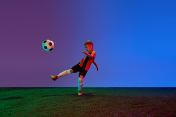 Male beginner soccer player in sports uniform in action, motion with ball over dark blue background in neon. Concept of sport, health, action, ad