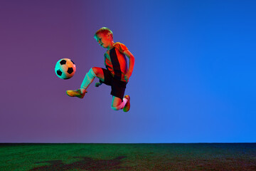 Fototapeta na wymiar Studio shot of little boy, soccer player in sports uniform jumping with football ball over dark blue background in neon. Concept of sport, health, action, ad
