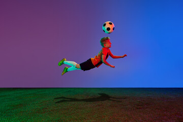 Active, energetic school age boy, football player training with soccer ball over dark blue background in neon. Concept of team sport, health, action, ad