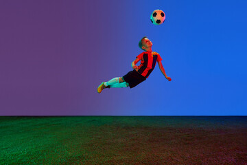 Studio shot of little boy, soccer player in sports uniform jumping with football ball over dark blue background in neon. Concept of sport, health, action, ad