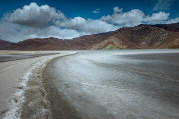 A walkway amidst the fully frozen Pangong Lake, inviting you to explore the captivating natural landscape on a sunny day, under a clear blue sky adorned with fluffy white clouds