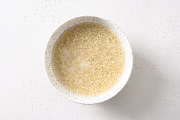 Making vegan milk of green buckwheat. Soaking process grain with water in white bowl on white background. Close up. Copy space.