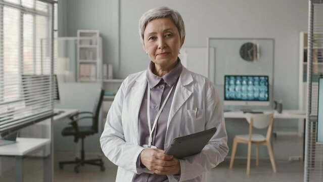 Medium portrait of mature Asian female doctor in white lab coat posing for camera with digital tablet in hands in medical clinic