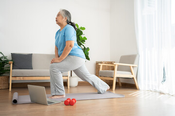 Senior Asian woman watching online courses on a laptop while exercising in the living room at home. Old woman stretching exercises in the living room at home, Concept of workout training online.