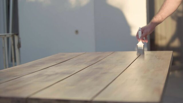 Male Caucasian hand applying primer on wood as unrecognizable man entering pointing at furniture. Process of wooden table painting outdoors on sunny day