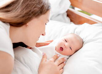 Young mother looking at her awaking newborn daughter swaddled in white sheets at home and lying with mouth open.