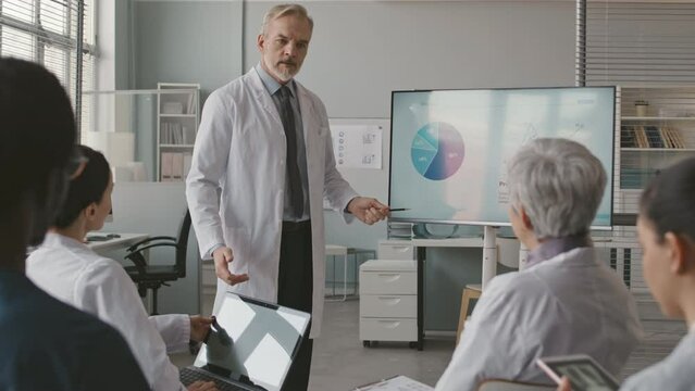 Medium shot of middle aged Caucasian male doctor standing by projector with slide presentation on it making speech to diverse team of colleagues listening to him during medical conference