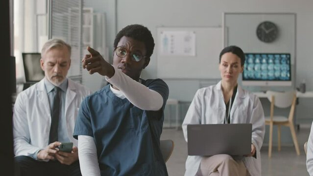 Medium shot of young African American male medical specialist wearing blue scrubs raising his hand to ask question during expert conference with other doctors sitting at their seats