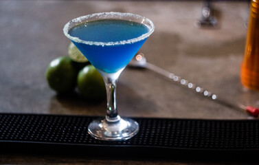 Summer alcohol fresh fruit drink. Blue colored cocktail with lemon on the bar counter has a copy space and a bar background.