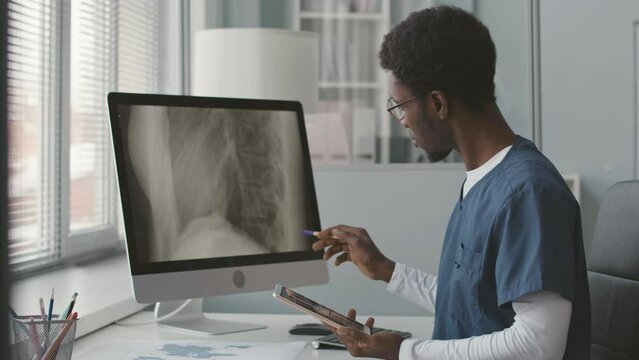 Waist up of young African American male doctor in blue scrubs looking at computer monitor while analyzing patient spine x-ray