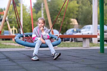 A teenage sad girl with long hair pulled back in a ponytail, wearing jeans, a hoodie and sneakers is sitting on a round swing on a sunny summer day.The concept of loneliness, problems of adolescence.