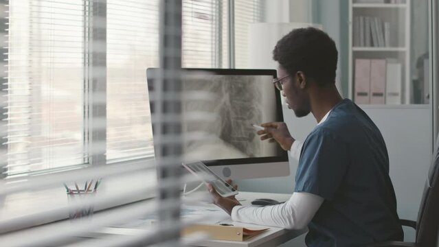 Waist up of young Black male physician sitting at desk in front of computer analyzing spine x-ray of patient on screen and conducting diagnosis
