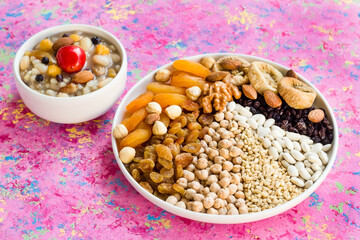 Uncooked Ashura ingredients chickpeas,wheat, rice and white beans,nuts and sweet fruits designed in white plate on pink surface with a bowl of Noah's Puding