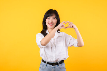 Young female healthcare. Asian woman wearing white shirt feels happy and romantic shapes heart gesture expresses tender feeling poses isolated on yellow background. People affection and care concept.