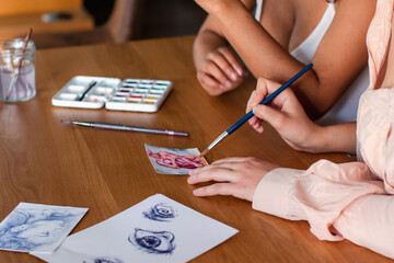 Side view of crop anonymous diverse lesbian couple drawing with paintbrushes on paper on wooden table